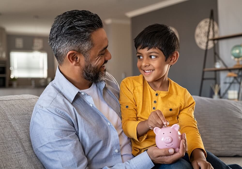 Indian father and smiling son putting coin into piggy bank. Smiling boy sitting on father lap saving money in piggybank. Middle eastern dad teaching son to save money while putting coin in piggy bank.